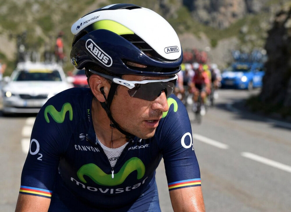 Osoba w kasku rowerowym z logo \'ABUS\' i \'Movistar\' jedzie na rowerze w wyścigu. W tle widoczne są samochody i inni kolarze na drodze.