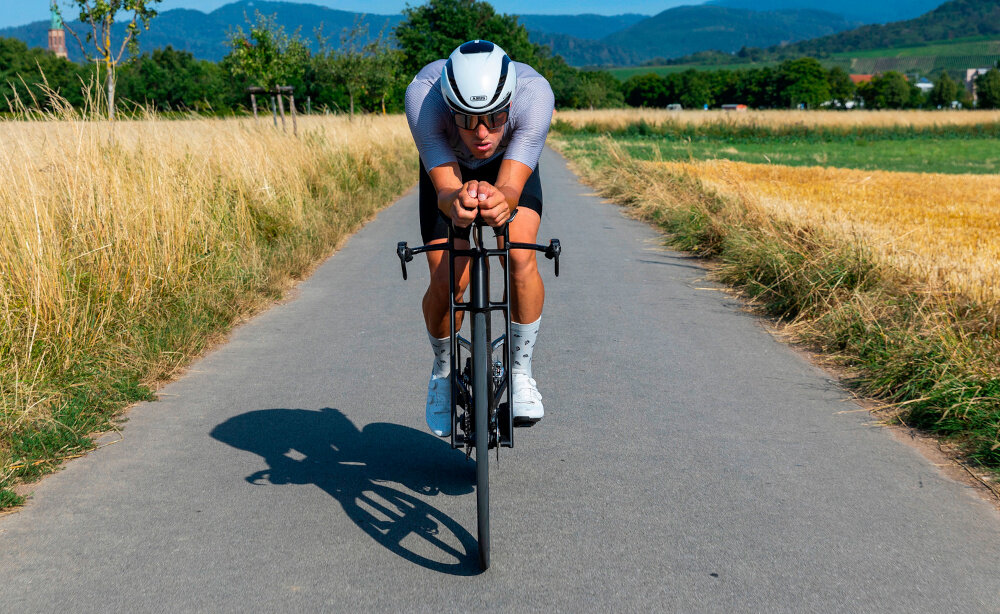 Kask rowerowy ABUS GameChanger 2.0 Shine Biały Szosowy rozmiar M, 57-61 cm MIPS osoba w kasku jedzie na rowrze po szosie w tle łąka pola bezpieczeństwo aerodynamiczna jazda