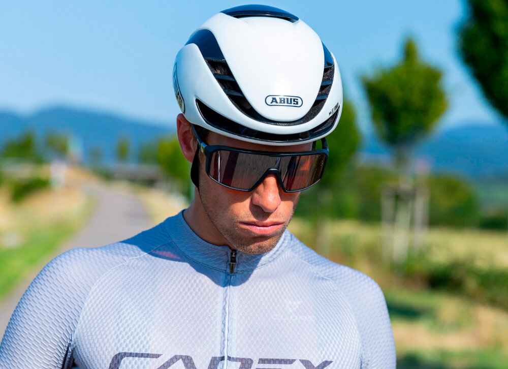 Kask rowerowy ABUS GameChanger 2.0 Shine Biały Szosowy rozmiar M, 57-61 cm MIPS krajobraz osoba w kasku na tle gór i łąki ActiCage piankowe wzmocnienie strukturalne proces In-Mold amortyzujący materiał EPS zewnętrzna powłoka z poliwęglanu dolna krawędź z PC