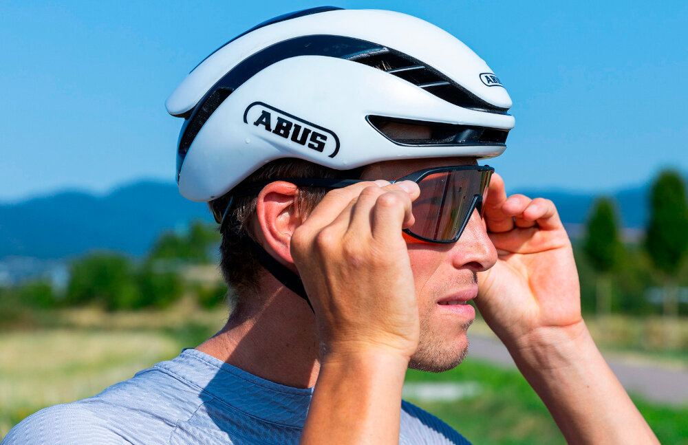 Kask rowerowy ABUS GameChanger 2.0 Shine Biały Szosowy rozmiar L, 57-61 cm MIPS krajobraz górski osoba zakłąda okulary osoba w kasku paski Flow Straps Pro ogranicza opór przepływu i hałas cienka górna część zapięcie magnetyczne FIDLOCK miękkie poduszki mają mocowanie na rzepach przedni i tylny uchwyt na okulary