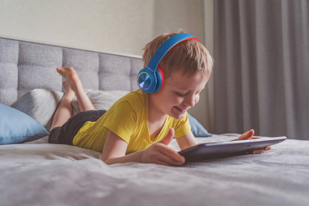 słuchawki nauszne POWERLOCUS Pled - Łączność Bluetooth, dziecko leżące na łóżku z założonymi słuchawkami i trzymające tableta w ręku