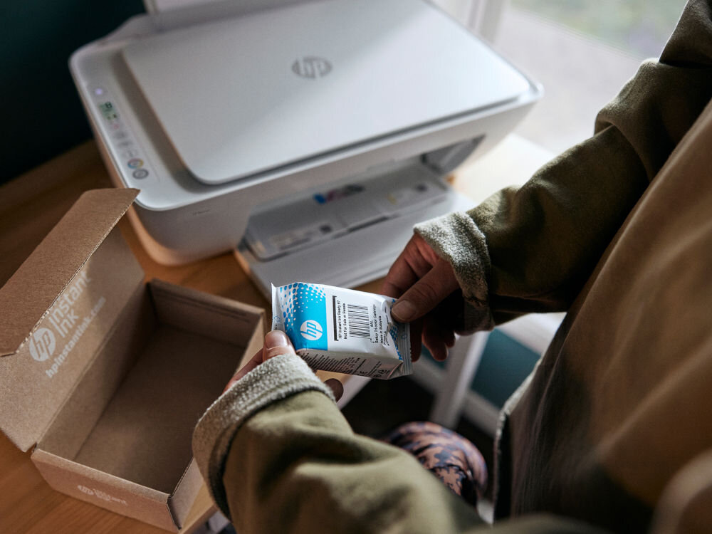 Urządzenie wielofunkcyjne HP DeskJet 2821e Druk w kolorze, Wi-Fi, Instant Ink prezentacja drukarki na biurku obok osoba trzymająca tusz świeżo wyjęty z kartonu dynamiczne zabezpieczenia wkładów sprawne działanie regenerowane wkłady powtórne napełnianie oszczędność przy eksploatacji bez rezygnacji z jakości