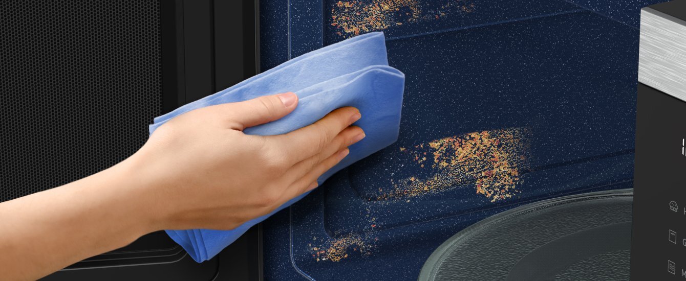 Osoba czyści wnętrze kuchenki mikrofalowej niebieską ściereczką. Wnętrze kuchenki jest zabrudzone resztkami jedzenia.