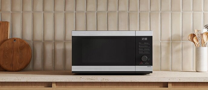 Na blacie kuchennym stoi czarno-srebrna kuchenka mikrofalowa z wyświetlaczem i przyciskami po prawej stronie. W tle widoczna jest ściana z jasnych płytek oraz drewniane akcesoria kuchenne.