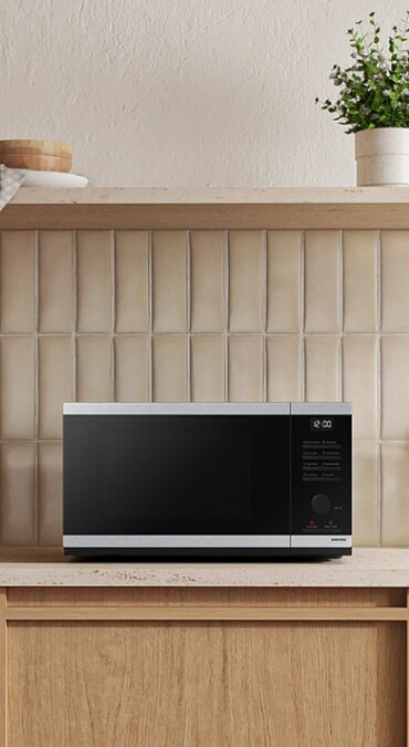 Czarno-srebrna kuchenka mikrofalowa stoi na blacie kuchennym przed jasnymi płytkami. Na wyświetlaczu widoczna jest godzina 12:00.