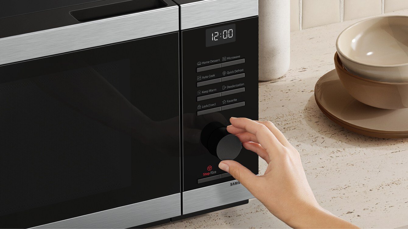 Osoba ustawia pokrętło na czarno-srebrnej kuchence mikrofalowej z wyświetlaczem pokazującym godzinę 12:00. Na panelu widoczne są przyciski z funkcjami: \'Home Dessert\', \'Microwave\', \'Auto Cook\', \'Quick Defrost\', \'Keep Warm\', \'Deodorization\', \'Lock/Unlock\', \'Favorite\'.