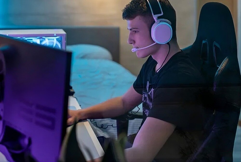 Router NETGEAR Nighthawk RS300 Młody mężczyzna w słuchawkach z mikrofonem siedzi w fotelu gamingowym przed monitorem w tle podświetlana obudowa komputera, prędkość, wydajność