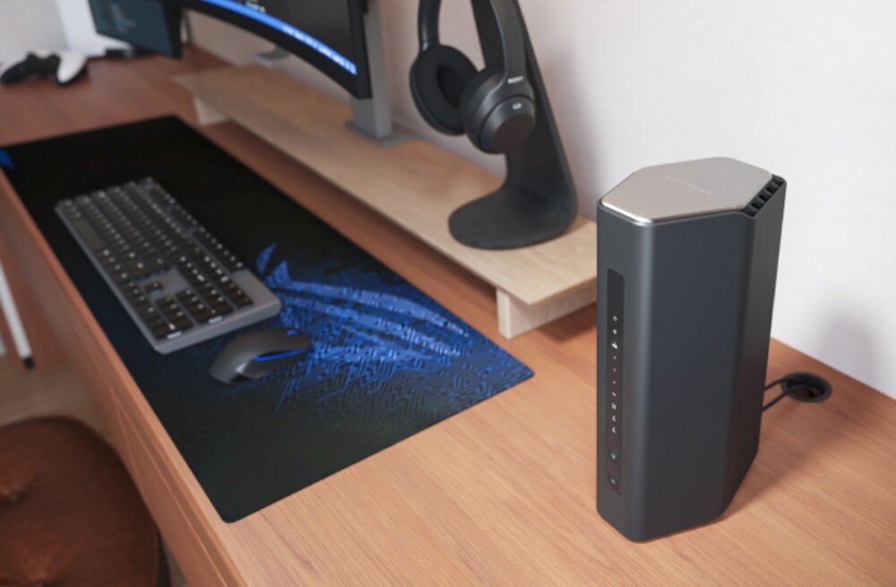Na biurku znajduje się czarny router o nowoczesnym designie z kilkoma diodami na przednim panelu. Obok routera widać klawiaturę i myszkę na dużej podkładce oraz słuchawki na stojaku.
