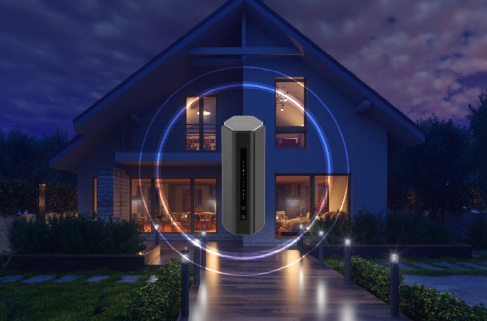 Na tle oświetlonego domu w nocy widoczny jest router z okręgami symbolizującymi zasięg sygnału. Router ma ciemny kolor i prostokątny kształt.