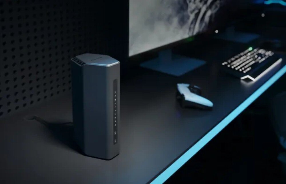 Na biurku znajduje się czarny router o nowoczesnym designie, obok którego leży biały kontroler do gier. W tle widoczny jest monitor i klawiatura z podświetleniem.