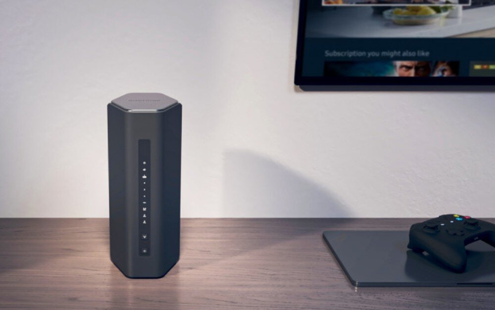 Na drewnianym biurku stoi czarny, sześciokątny router z pionowym panelem kontrolnym. Obok leży czarny kontroler do gier i laptop, a na ścianie wisi ekran z widocznym interfejsem użytkownika.