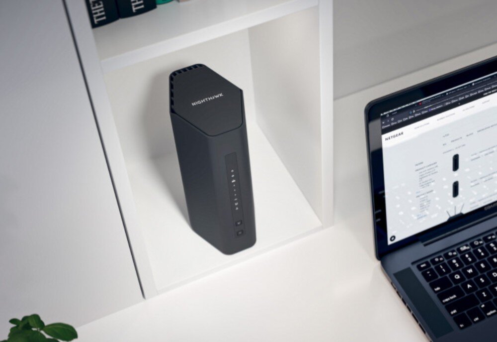 Router NETGEAR Nighthawk RS200 Pionowy router w kształcie graniastosłupa stoi na białej półce obok otwartego laptopa, na którego ekranie widać stronę internetową, opis