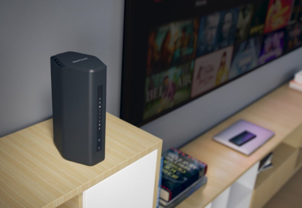 Router NETGEAR Nighthawk RS200 Pionowy router stoi na jasnym, drewnianym meblu w salonie, w tle widać fragment dużego ekranu telewizora z menu wyboru filmów, zabezpieczenia, firewall, szyfrowanie