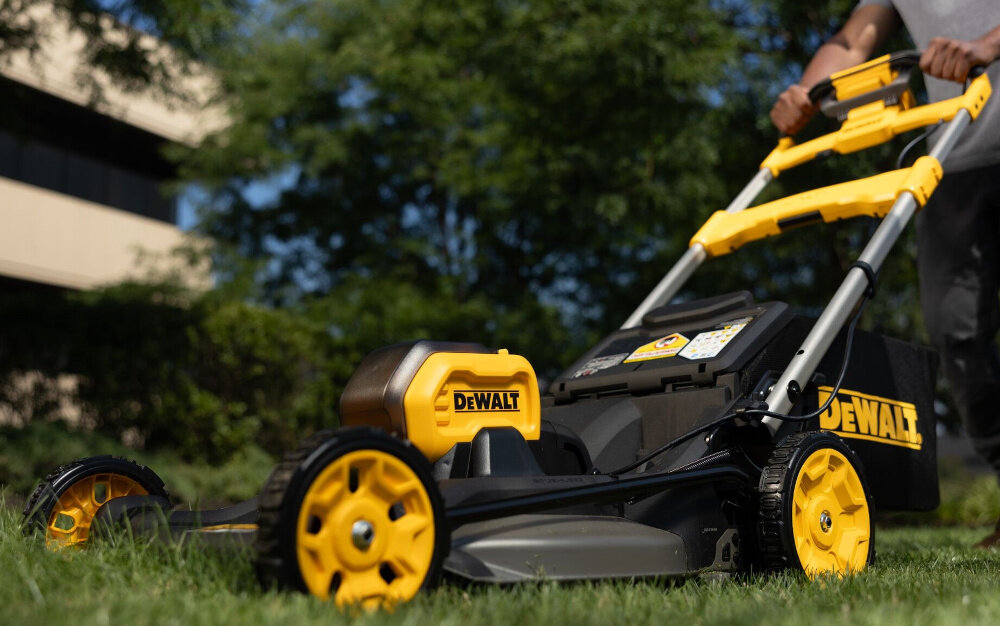 Kosiarka akumulatorowa DEWALT DCMWSP550N-XJ z napędem ogród trawnik osoba kosi trawę w tle drzewa budynek niebo napęd na tylne koła regulowana prędkość jazdy zmniejsza zmęczenie poprawia komfort pracy