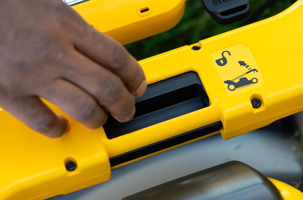 Kosiarka akumulatorowa DEWALT DCMWSP550N-XJ z napędem zbliżenie na przycisk służący do złożenia kosiarki osoba zbliża dłoń do przycisku waga około 29 kg składany uchwyt obsługiwany jednym przyciskiem lepsza organizacja przestrzeni