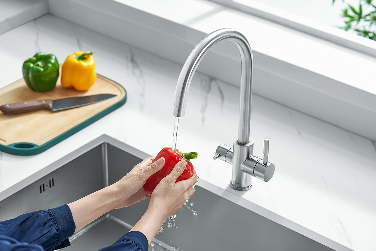 Osoba myje czerwoną paprykę pod strumieniem wody z metalowej baterii kuchennej. Na blacie obok znajduje się deska do krojenia z zieloną i żółtą papryką oraz nożem.