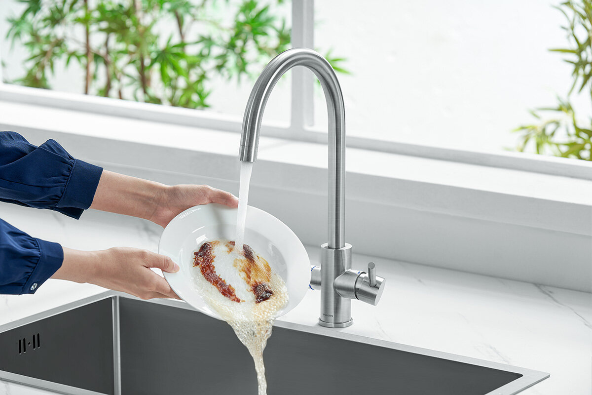 Osoba myje talerz pod strumieniem wody z metalowej baterii kuchennej o srebrnym wykończeniu. W tle widoczne są zielone rośliny za oknem.