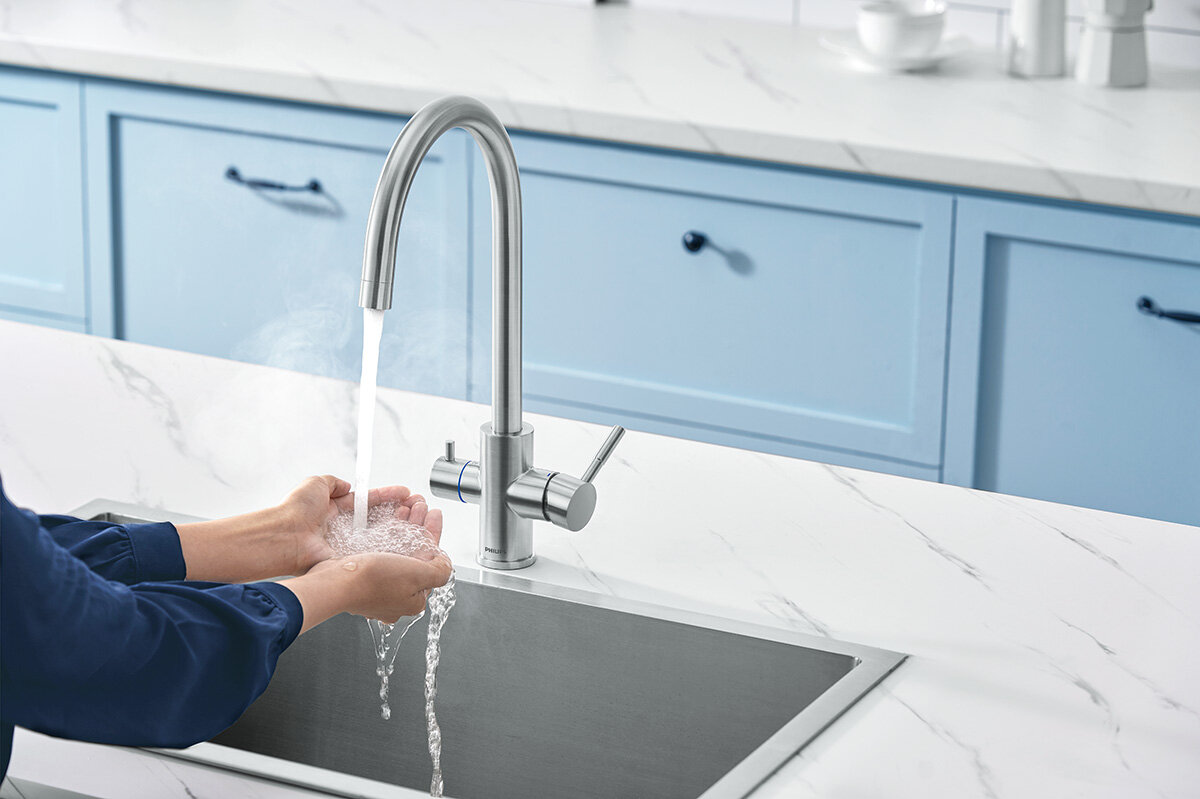 Osoba myje ręce pod strumieniem wody z baterii kuchennej o srebrnym, szczotkowanym wykończeniu. W tle widoczna jest jasna kuchnia z niebieskimi szafkami i białym blatem.