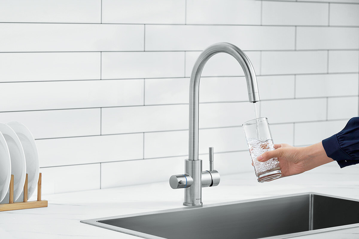 Na obrazie widoczna jest srebrna bateria kuchenna z wysokim, zakrzywionym wylewem, z której nalewana jest woda do szklanki trzymanej przez osobę. W tle znajduje się biała ściana z płytek oraz stojak z talerzami.