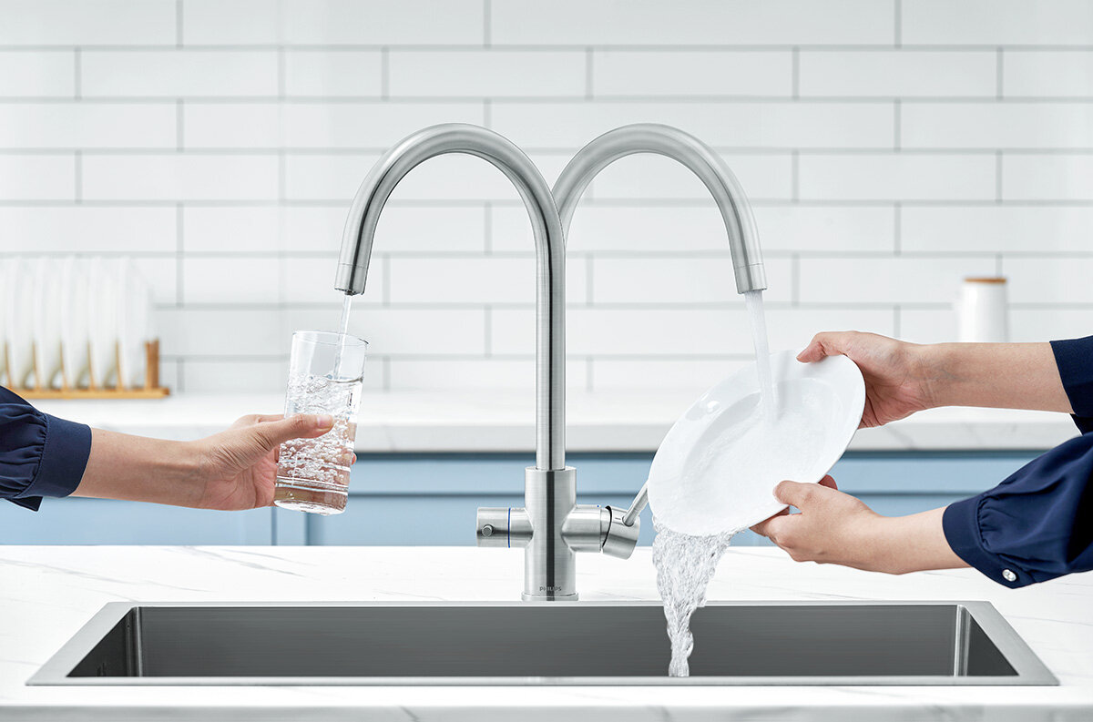 Na obrazie widoczna jest trójdrożna bateria kuchenna w kolorze szczotkowanego srebra, z której leci woda do szklanki i na talerz. W tle znajduje się biała ściana z kafelkami i niebieskie szafki.