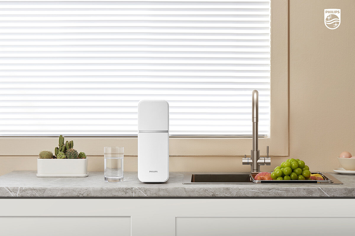Na blacie kuchennym znajduje się biały pojemnik z napisem \'PHILIPS\', szklanka z wodą, doniczka z kaktusami oraz bateria kuchenna w kolorze srebrnym. W tle widoczne są żaluzje na oknie i logo \'PHILIPS\' w prawym górnym rogu.
