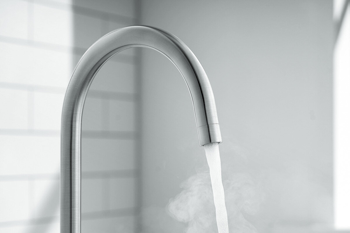 Na obrazie widoczna jest bateria kuchenna w kolorze szczotkowanego srebra, z której płynie woda. W tle znajduje się rozmyta ściana z jasnymi płytkami.
