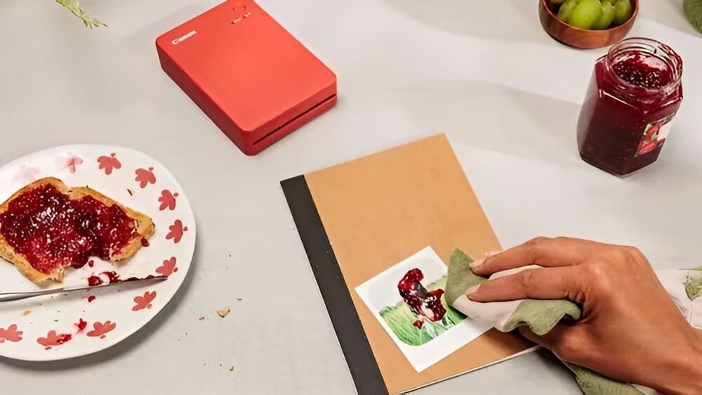 Drukarka CANON Selphy Square QX20 Druk w kolorze, Wi-Fi Szary urządzenie na jasnym stole obok talerz z kanapką naklejanie naklejki na zeszyt odporność na upływ czasu trwałość materiałów kolory nie blakną