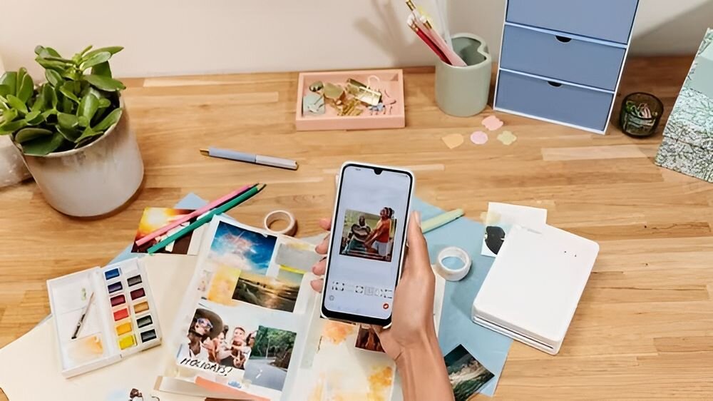 Drukarka CANON Selphy Square QX20 Druk w kolorze, Wi-Fi Szary urządzenie położone na stole wśród kartek zeszytów i rzeczy biurowym ręka trzymająca telefon Wi-Fi Direct Wi-Fi połączenie smartfona z drukarką bez routera komfort użytkowania mobilny charakter sprzętu