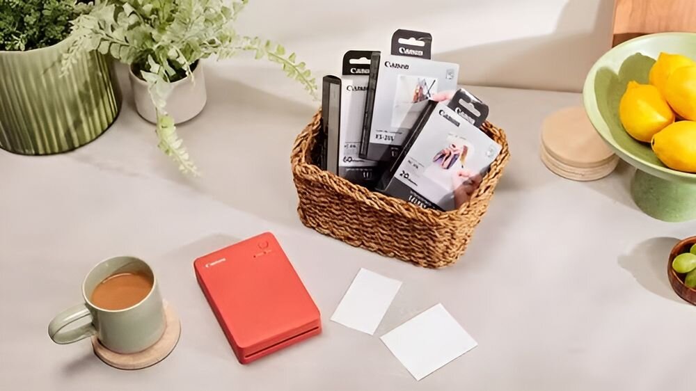 Drukarka CANON Selphy Square QX20 Druk w kolorze, Wi-Fi Czerwony drukarka położona na stole obok rozmiary papieru kubek rośliny w doniczkach w koszyku opakowania z kasetami i papierem mobilność dwa rozmiary dopasowanie do bieżących potrzeb