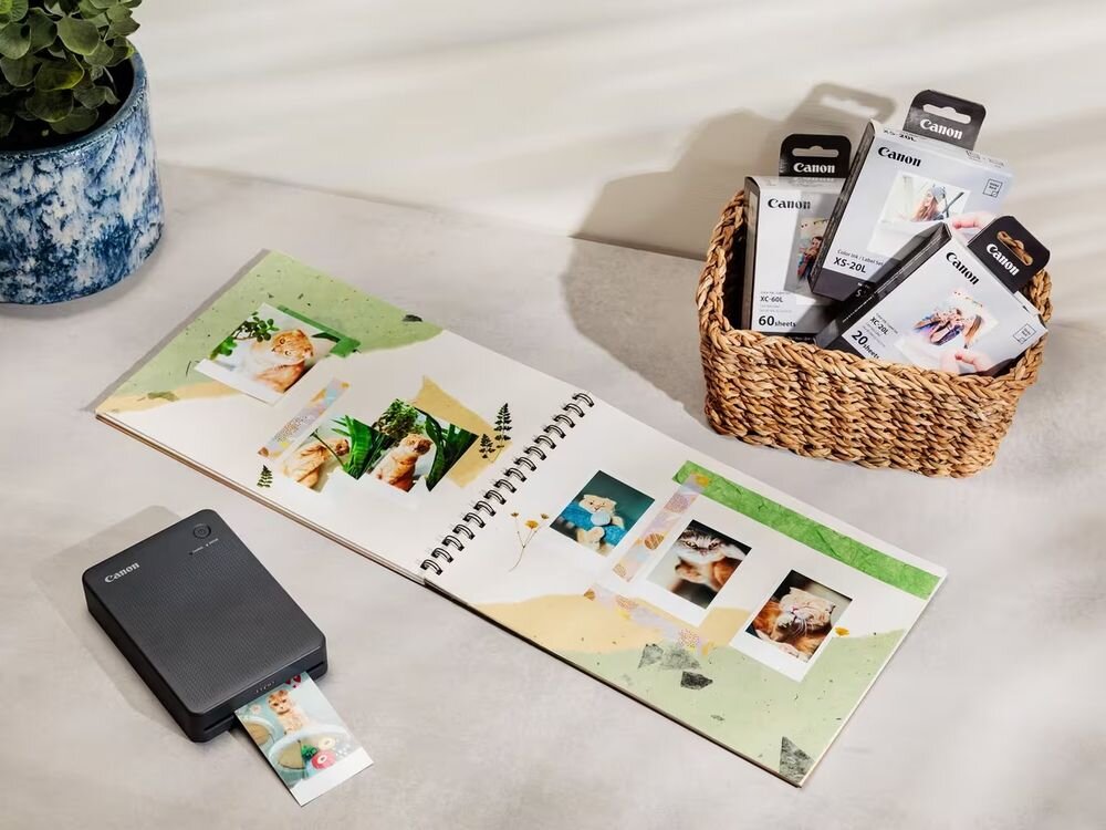 Drukarka CANON Selphy Square QX20 Druk w kolorze, Wi-Fi Czerwony drukarka na jasnym tle podczas drukowania obok album ze zdjęciami i koszyk z atramentem i papierem druk termosublimacyjny trzy cykle przenoszenia bogate obrazy w kolorze 256 poziomów na każdą barwę 24-bit warstwa ochronna