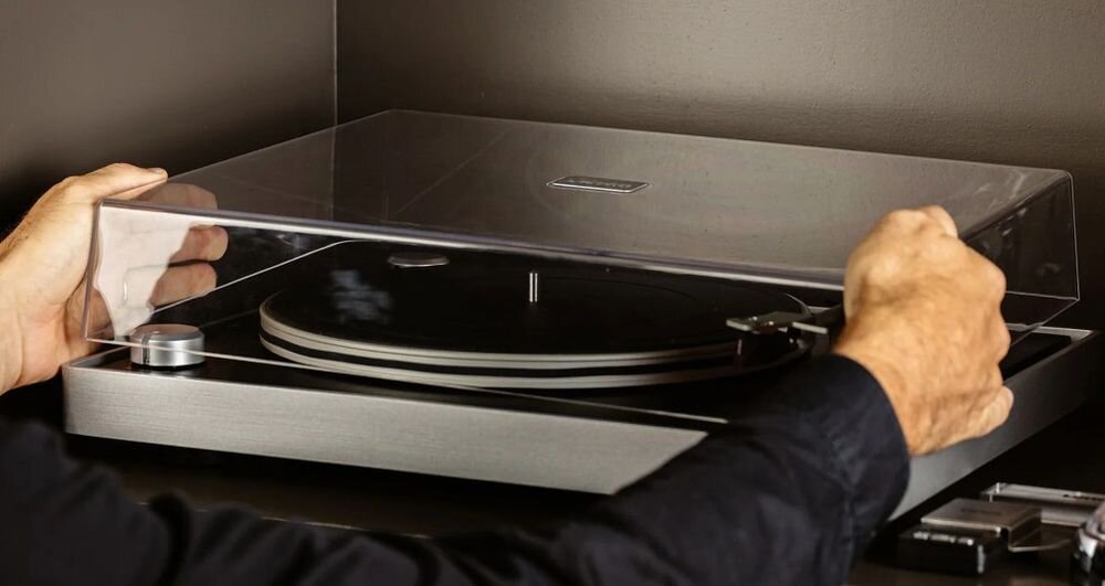 Gramofon LENCO L-455BK widoczny napęd paskowy 33 45 RPM stabilne obroty, gramofon stojący na półce w pomieszczeniu, osoba otwierająca pokrywe gramofonu