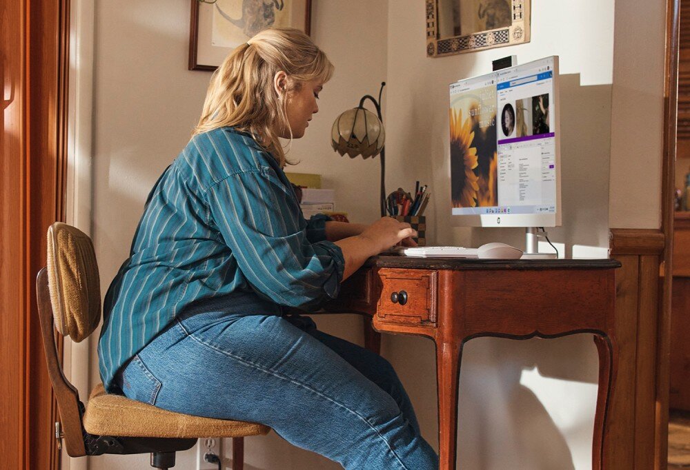 Komputer HP All-in-One 27-cr0222nw Osoba siedząca przy biurku i pracująca na komputerze z dużym monitorem ustawionym na drewnianym stole w jasnym pomieszczeniu, opis