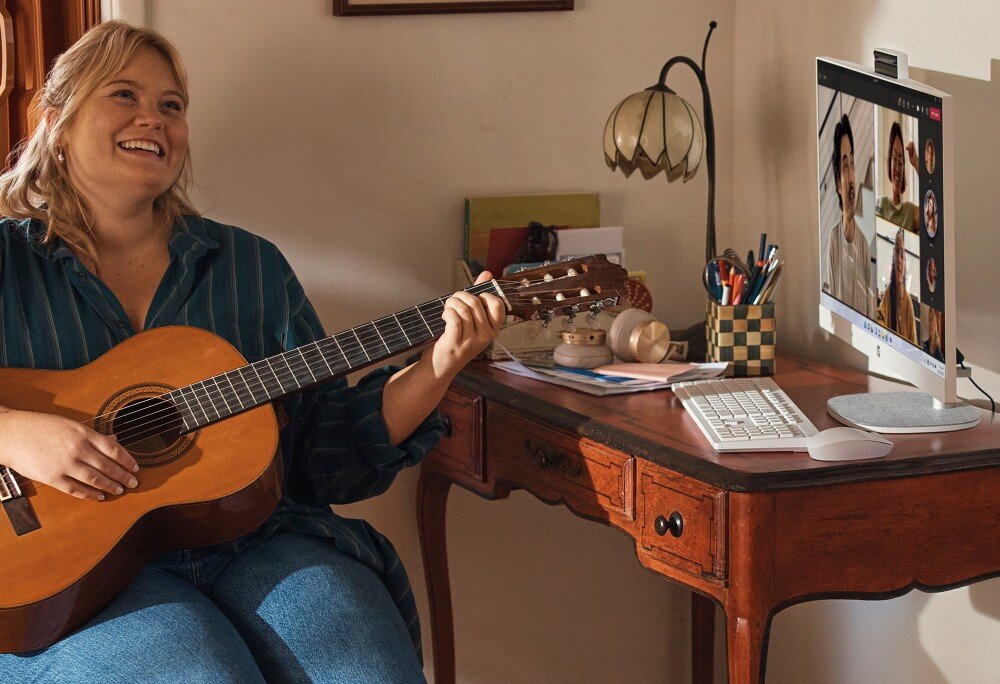 Komputer HP All-in-One 27-cr0222nw Osoba siedząca z gitarą i grająca przed komputerem, na którego ekranie widoczna jest wideokonferencja z kilkoma uczestnikami, kamera internetowa, mikrofon