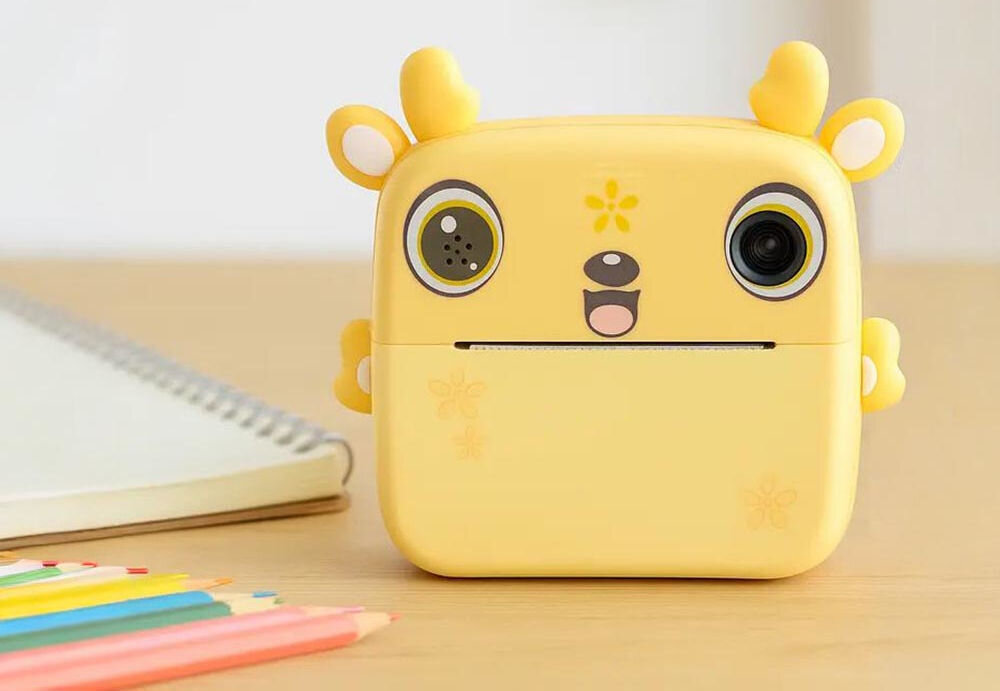 Drukarka EXTRALINK EPP-006 Złącze USB Żółty + Papier termiczny zwykły (3 szt.) aparat natychmiastowy stojący na drewnianym biurku obok kolorowych kredek i notesu druk termiczny czarno-biały obraz