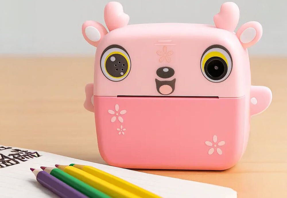 Drukarka EXTRALINK EPP-005 Złącze USB Różowy + Papier termiczny zwykły (3 szt.) aparat natychmiastowy stojący na drewnianym biurku obok kolorowych kredek i notesu druk termiczny czarno-biały obraz