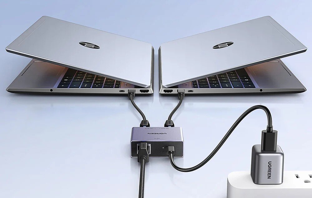 Splitter UGREEN NW301 45311 Dwa laptopy ustawione obok siebie, połączone kablami do małego, szarego urządzenia rozdzielającego sygnał, które jest dodatkowo zasilane z gniazdka ściennego