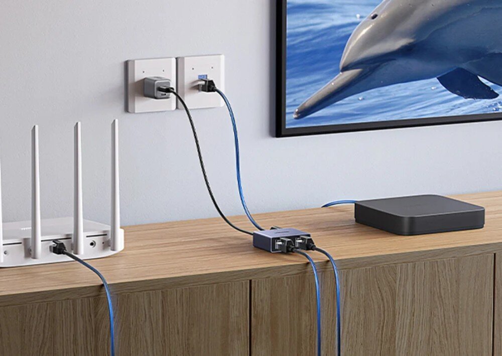 Splitter UGREEN NW301 45311 Urządzenie leżące na drewnianej komodzie w salonie, podłączone do routera, gniazdka ściennego oraz telewizora i przystawki smart tv za pomocą niebiesko-czarnych kabli