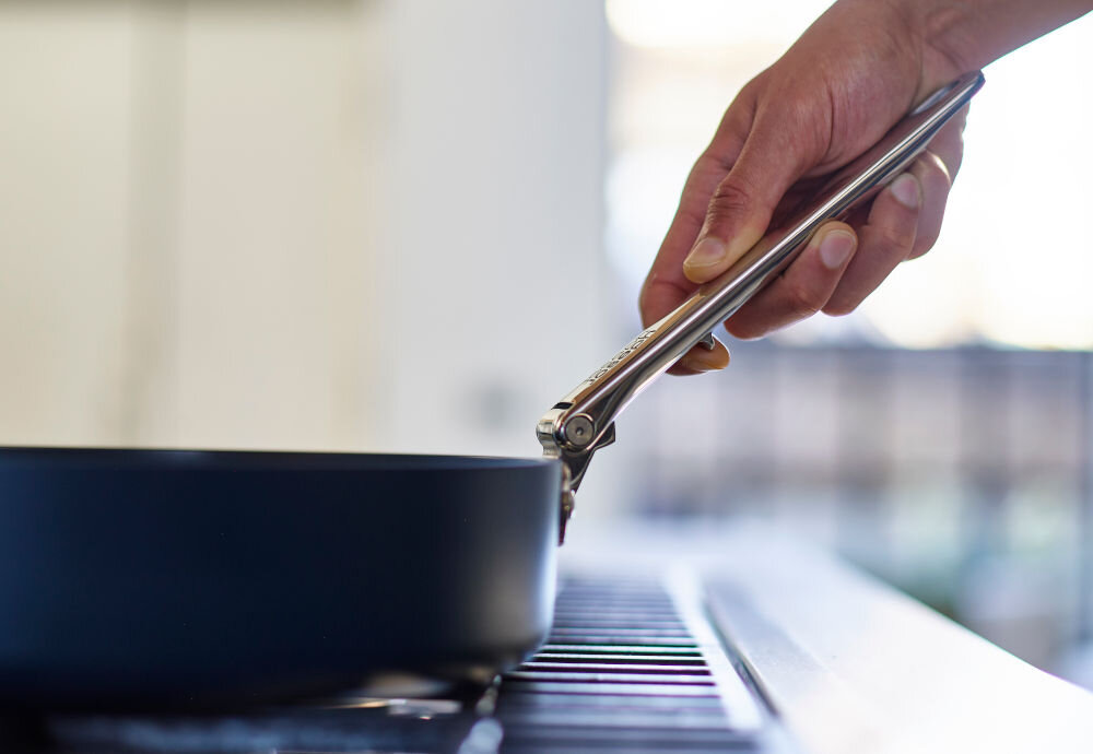 JJ-Zestaw 2patelni ze skł.uchwytami+przekład.Space ręka trzymająca srebrną rączkę patelnia na gazówce  składane uchwyty długi i przedni zajmują mniej miejsca do małych kuchni większa patelnia średnica 28 cm mniejsza 24 cm uchwyty z polerowanej stali nierdzewnej połączone z korpusami opatentowane mocowanie SwingLock