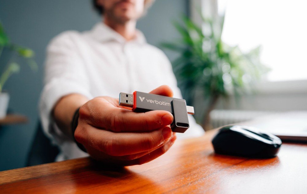 Pendrive VERBATIM Dual QuickStick Osoba siedząca przy biurku trzymająca w palcach szary pendrive skierowany przodem w stronę obiektywu