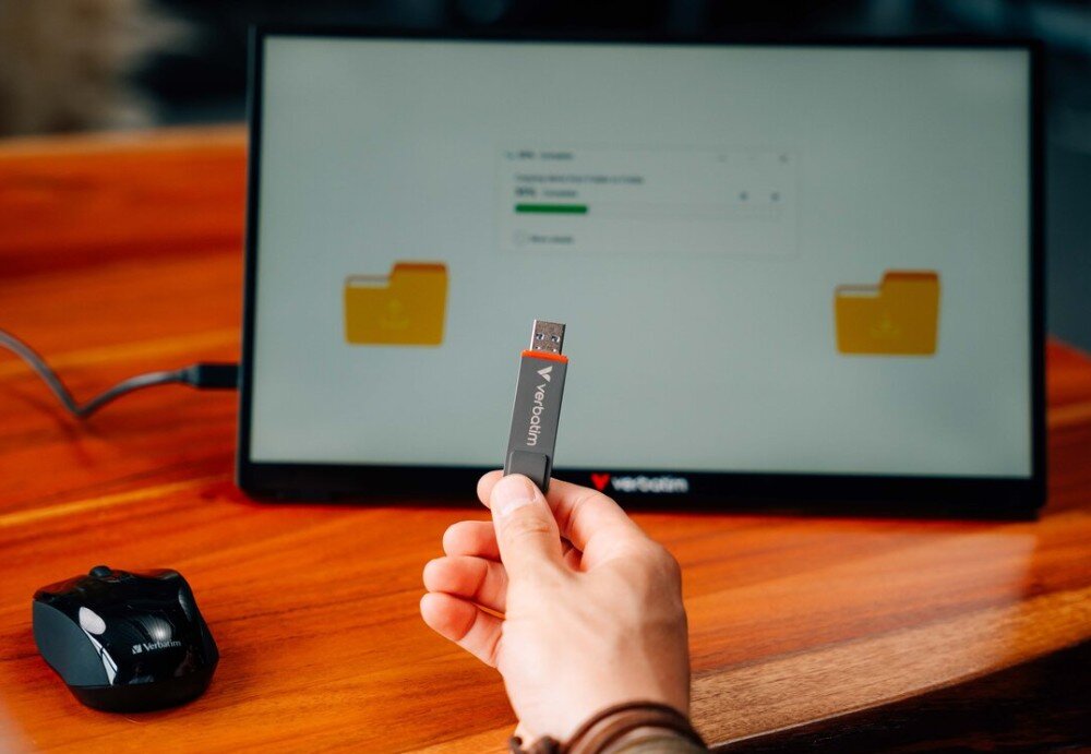 Pendrive VERBATIM Dual QuickStick Pamięć przenośna podłączona do portu tabletu trzymanego oburącz nad powierzchnią drewnianego blatu