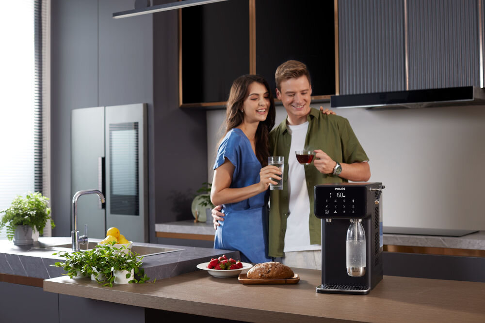 Dyspenser do wody PHILIPS ADD5962BK/10 Uśmiechnięta para w kuchni trzymająca szklankę wody i filiżankę herbaty obok wielofunkcyjnego dystrybutora wody Philips.