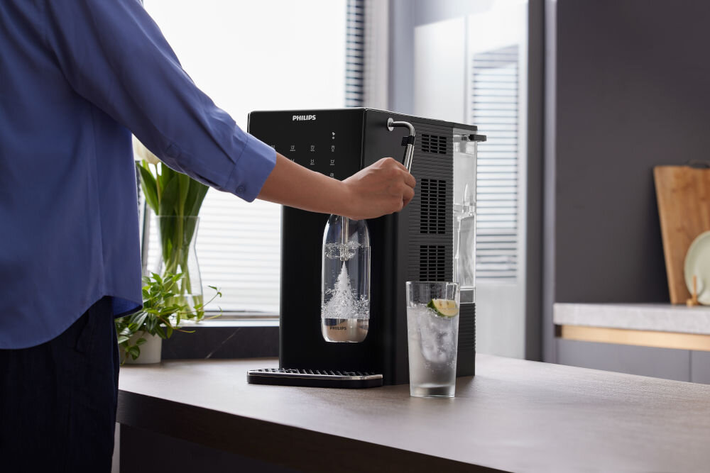 Dyspenser do wody PHILIPS ADD5962BK/10 Osoba przygotowująca wodę gazowaną za pomocą bocznej dźwigni saturatora w dystrybutorze Philips; obok szklanka wody z lodem i cytryną.