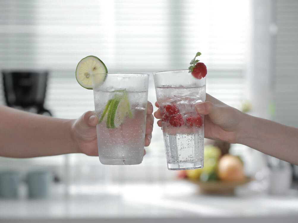 Dwie szklanki z wodą gazowaną, w jednej plasterki limonki, w drugiej maliny. W tle rozmyta kuchnia z owocami na blacie.