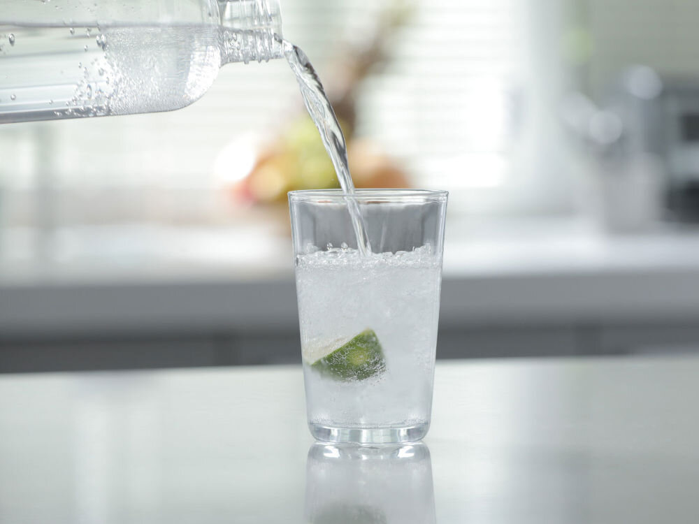 Na obrazie widać szklankę z wodą gazowaną i kawałkiem limonki, do której nalewana jest woda z butelki. W tle znajduje się rozmyte wnętrze kuchni.