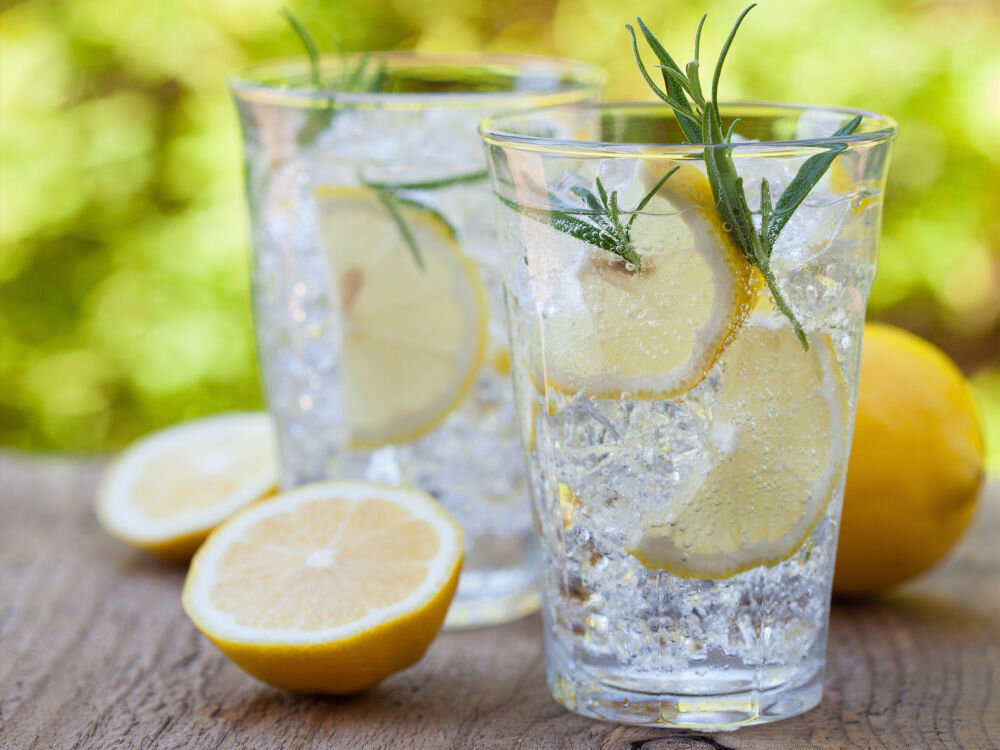 Na drewnianym stole stoją dwie szklanki z napojem, w których znajdują się plasterki cytryny i gałązki ziół. Obok leży przekrojona cytryna i cała cytryna.