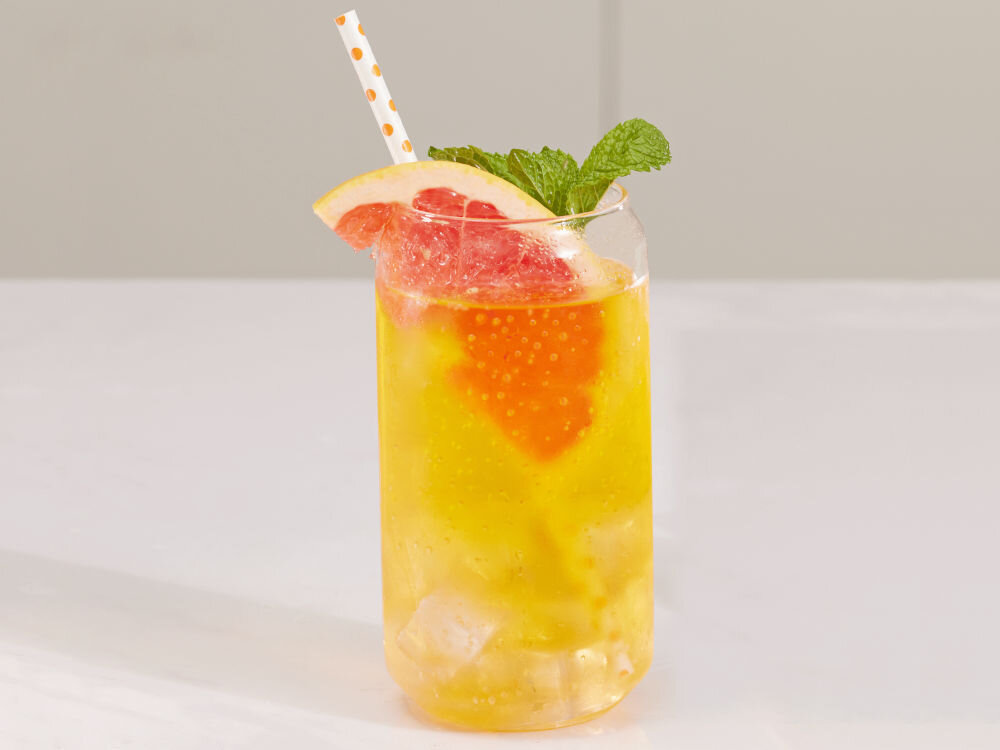 Szklanka z żółtym napojem, kostkami lodu, plasterkiem grejpfruta i listkiem mięty. W szklance znajduje się słomka w kropki.