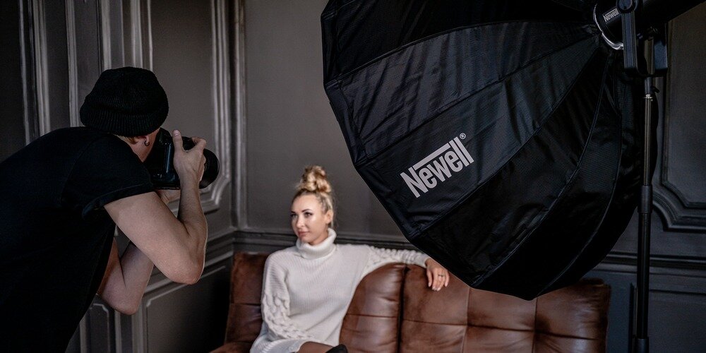 Fotograf wykonuje zdjęcie modelce siedzącej na kanapie, oświetlonej parabolicznym softboxem Newell 120 cm
