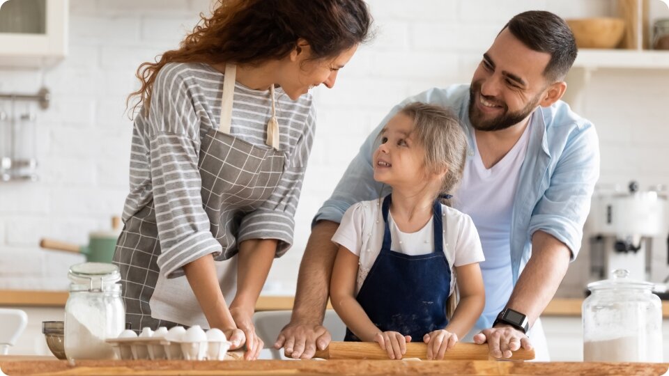 Uśmiechnięta rodzina – mama, tata i córka w fartuchach – wspólnie przygotowuje ciasto w jasnej, nowoczesnej kuchni