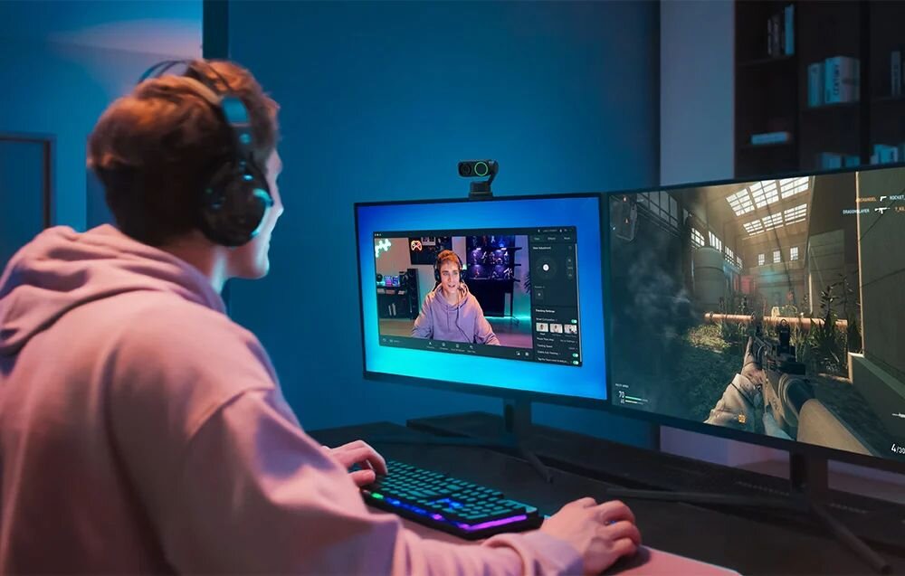 INSTA360 Link 2 Insta360 Link 2 1/2-calową matrycę, rozdzielczość Ultra HD 4K, Technologia HDR efekt bokeh rozmycie tła - Gracz w słuchawkach siedzący przed dwoma monitorami i kamerą internetową w oświetlonym na niebiesko pokoju