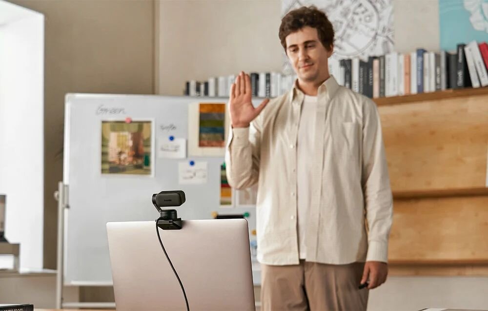 INSTA360 Link 2 Insta360 Link 2 aplikacji na smartfonie sterowanie gestami przybliżać i oddalać obraz, uruchamiać śledzenie AI funkcję wykrywania białej tablicy - Mężczyzna stojący przed otwartym laptopem z kamerą internetową i wykonujący gest dłonią